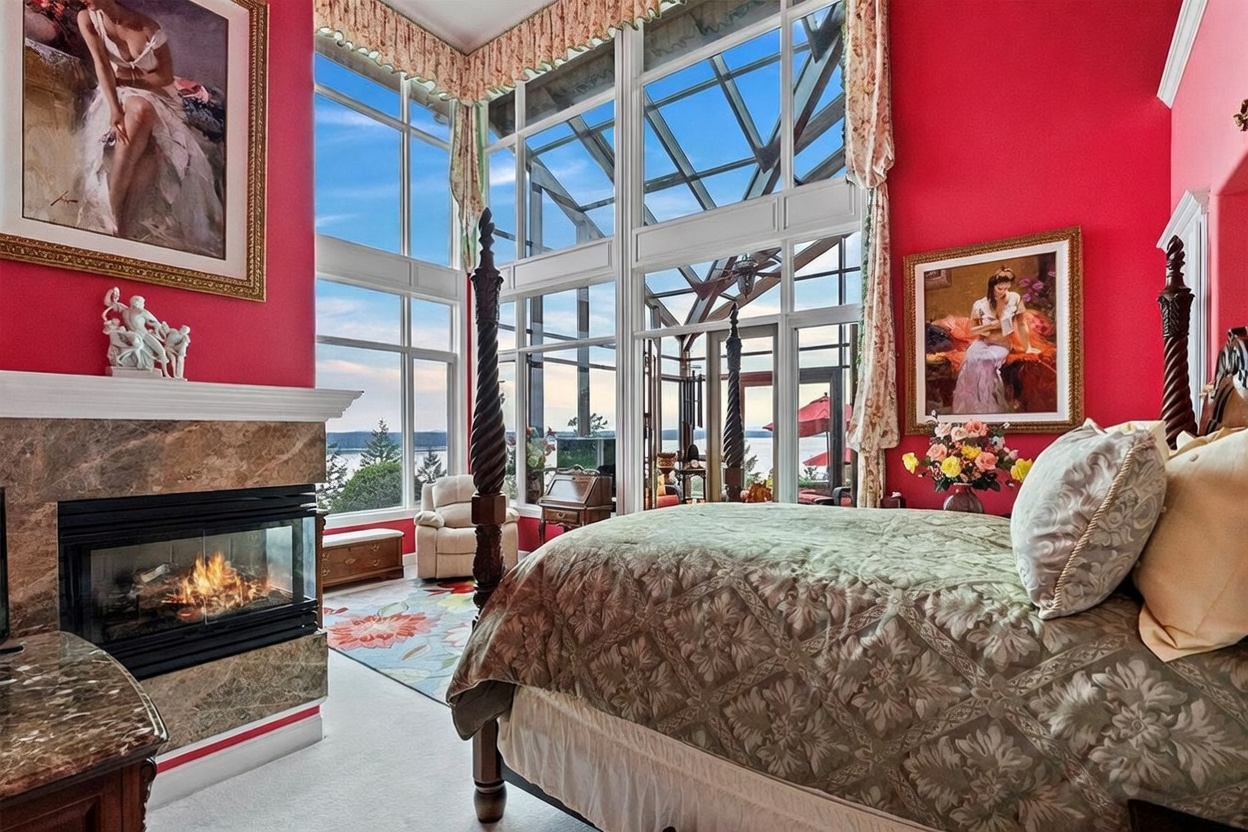 This primary bedroom features a dramatic, high-ceilinged design with expansive glass walls that offer panoramic water views. The room is anchored by a four-poster bed with a patterned comforter and a stone-clad fireplace surround, all set against vibrant red walls. The space is decorated with ornate artwork and traditional furnishings, creating a bold and luxurious atmosphere.