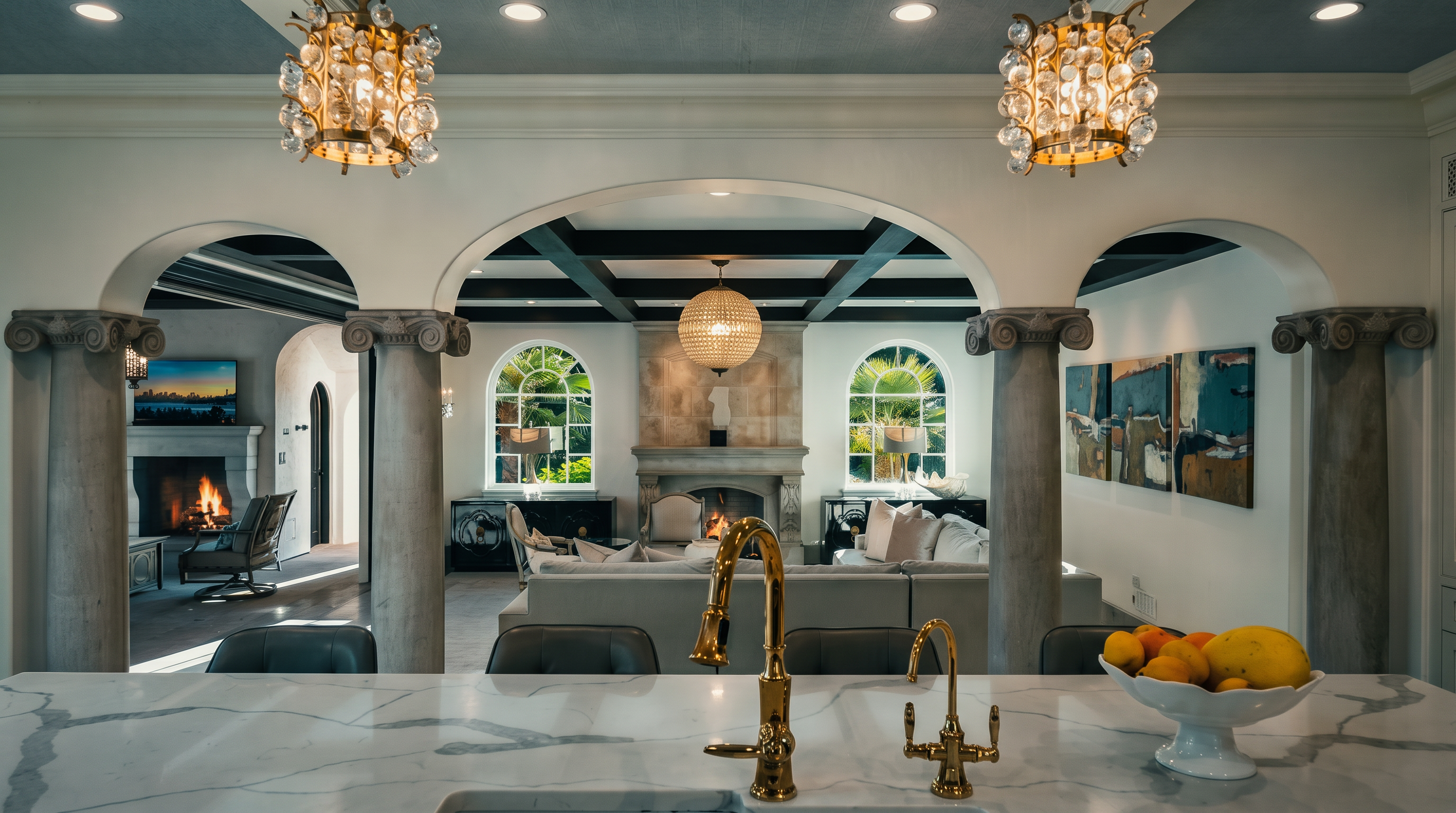This view from a kitchen island looks through a series of grand, arched openings supported by stone columns into a spacious living area. The foreground features a white stone countertop with a gold faucet and a bowl of fruit, while the background showcases a sophisticated living room with a stone fireplace, coffered ceilings, and large arched windows. The overall aesthetic is elegant and Mediterranean-inspired, emphasizing an open-concept layout that connects the culinary and relaxation spaces.