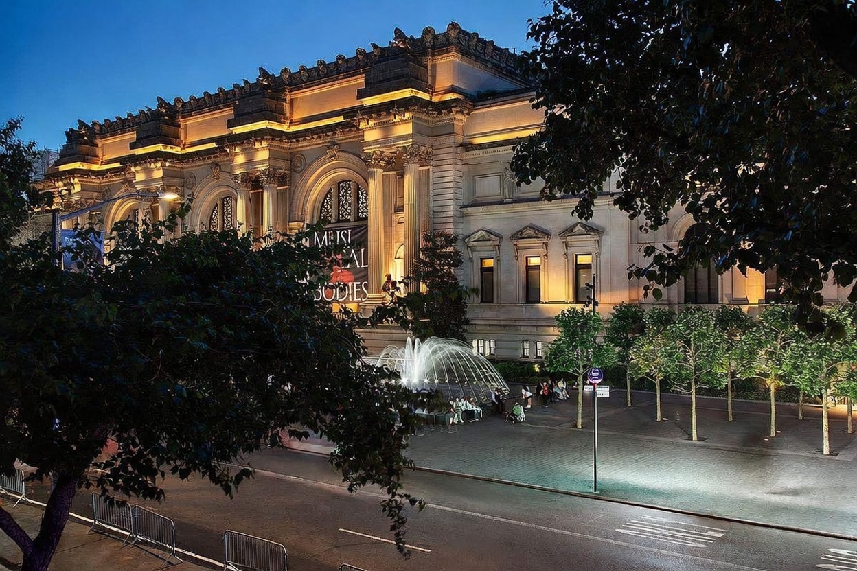 This cinematic, dusk-lit shot captures the iconic facade of The Metropolitan Museum of Art in New York City, showcasing its grand neoclassical architecture and illuminated stone columns. A vibrant fountain sprays water in the foreground, set against a paved plaza lined with trees and pedestrian walkways. The perspective is elevated, providing a sweeping view of the museum's impressive scale and the surrounding urban environment.