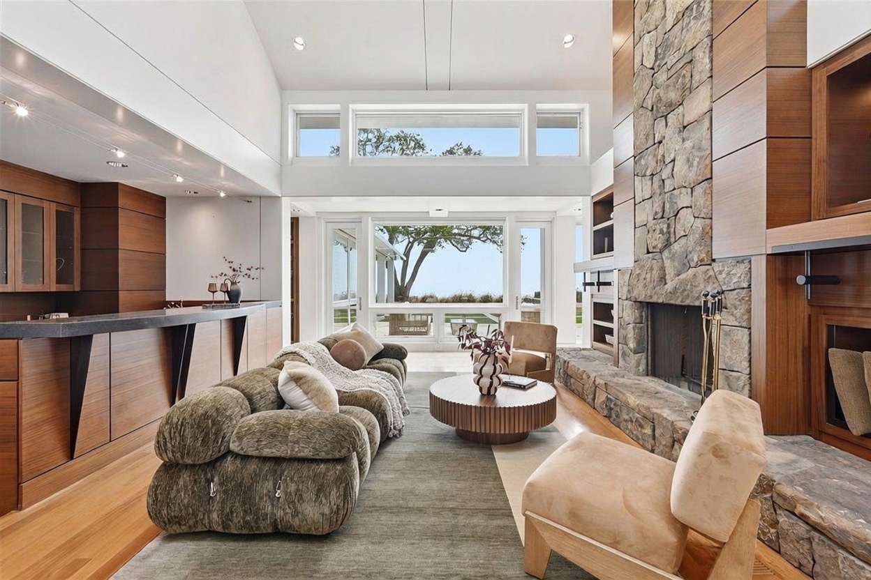 This spacious living room features a high, vaulted ceiling and a prominent stone-clad chimney breast that anchors the space. A large, textured sofa and a circular coffee table are arranged on a neutral area rug, while a pair of chairs provides additional seating near the expansive windows that offer a view of the outdoors. The room is finished with warm wood cabinetry and trim, creating a sophisticated and inviting atmosphere.