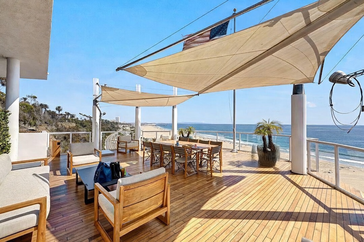 This expansive outdoor deck offers a luxurious, elevated vantage point overlooking the ocean and sandy coastline. The space is furnished with a large dining set and comfortable lounge seating, all shaded by modern fabric sails. The wooden decking and clear glass railings create a seamless, open-air atmosphere perfect for coastal entertaining.