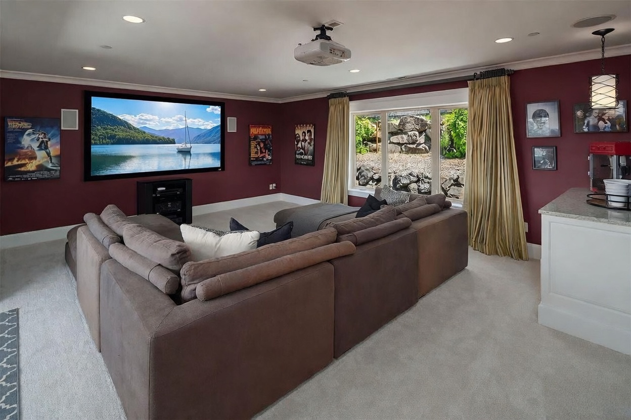 This dedicated home theater room features deep red walls, a large projection screen, and a comfortable, oversized sectional sofa arranged for optimal viewing. A ceiling-mounted projector and integrated wall speakers enhance the cinematic experience, while a small refreshment area with a popcorn machine adds a functional touch. Large windows with elegant curtains allow natural light to enter, providing a versatile space that transitions easily from a movie room to a casual lounge.