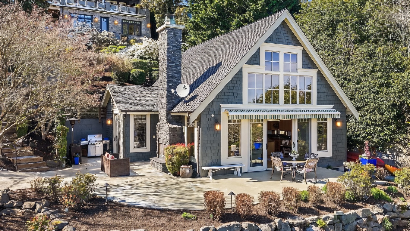 This charming, shingled cottage-style home features a prominent stone chimney and a spacious concrete patio perfect for outdoor entertaining. The rear facade showcases large windows and a glass-paned door that seamlessly connects the interior living space to the landscaped backyard. The setting is serene and picturesque, nestled against a lush, terraced hillside.