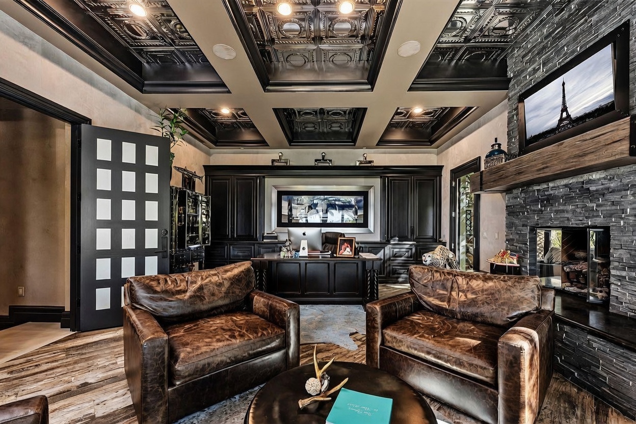 This sophisticated home office features a dramatic coffered ceiling with metallic-finished panels and dark trim, creating a luxurious and moody atmosphere. The room is anchored by a large, dark wood desk and two plush, distressed leather armchairs positioned around a circular coffee table. A prominent stone-clad chimney breast with a wooden mantel serves as a focal point, while built-in cabinetry provides extensive storage and a professional aesthetic.
