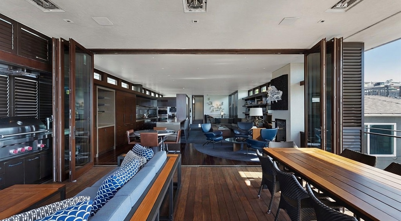 This expansive indoor-outdoor living space features a covered deck with a dining area and lounge seating, seamlessly connected to the interior living and kitchen areas through wide-opening folding glass doors. The interior showcases a contemporary open-concept design with dark wood flooring, a sleek kitchen, and a comfortable lounge area centered around a fireplace. The overall aesthetic is sophisticated and modern, emphasizing a fluid transition between the outdoor deck and the main living quarters.