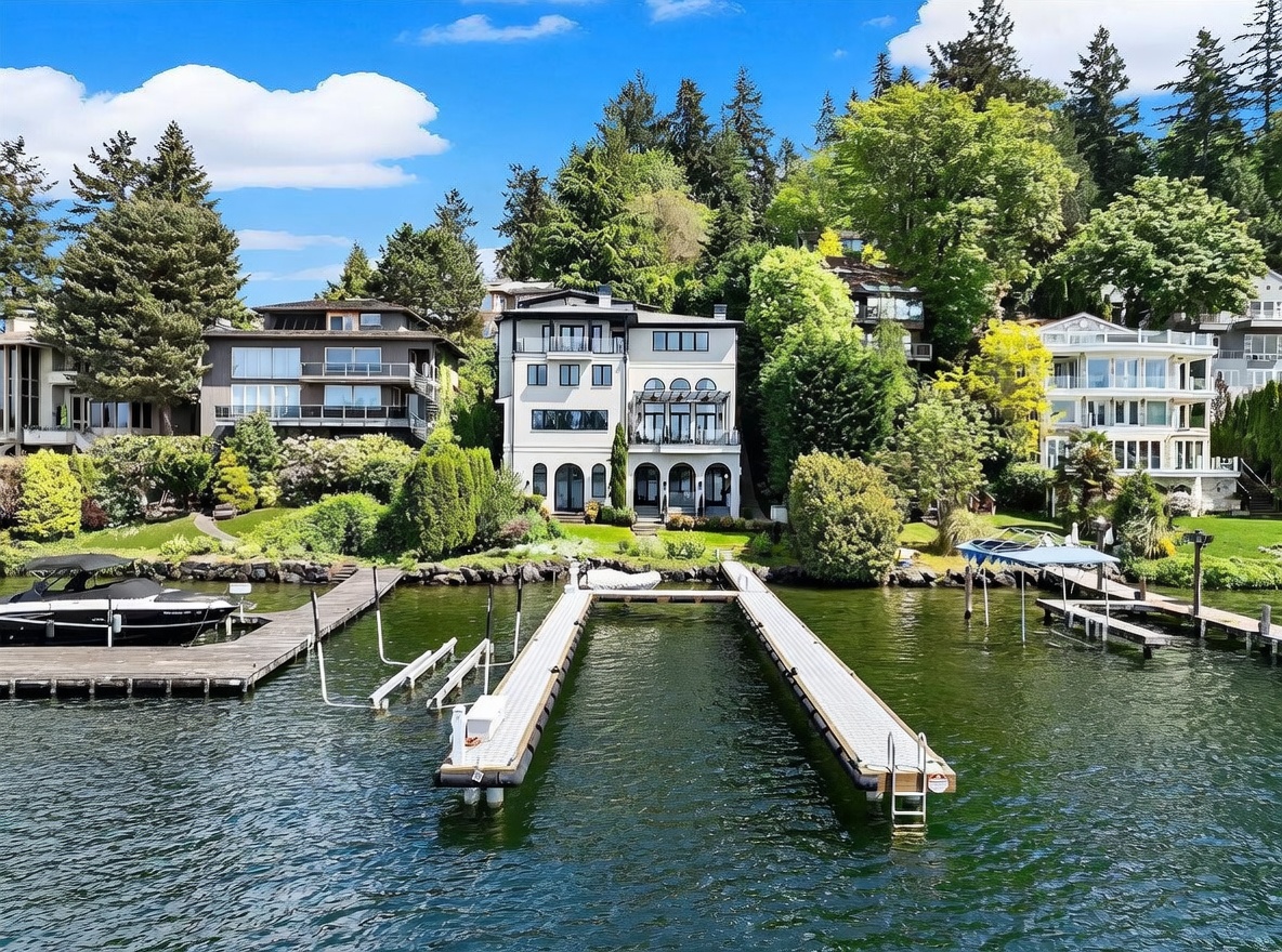 This waterfront property features a multi-story residence with a classic architectural style, characterized by symmetrical windows and a tiered balcony design. The home is nestled into a lush, tree-covered hillside and includes a private dock extending into the calm lake. The perspective is captured from the water, showcasing the home's prominent position and its direct access to the shoreline.