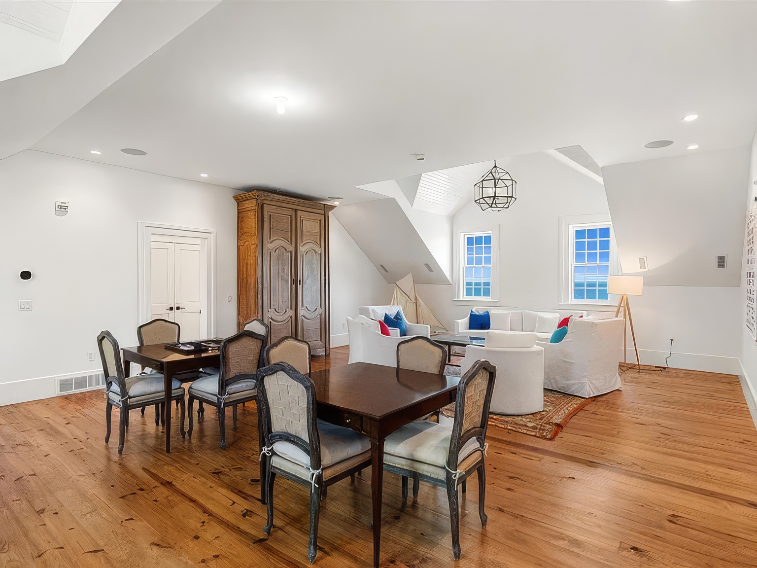 This spacious, open-concept room features a blend of dining and living areas under a vaulted ceiling with architectural dormers. The space is anchored by warm-toned wood flooring and includes a formal dining set, a large wooden armoire, and a comfortable seating area arranged around a central rug. Large windows provide ample natural light and views, creating an airy and inviting atmosphere suitable for both entertaining and relaxation.