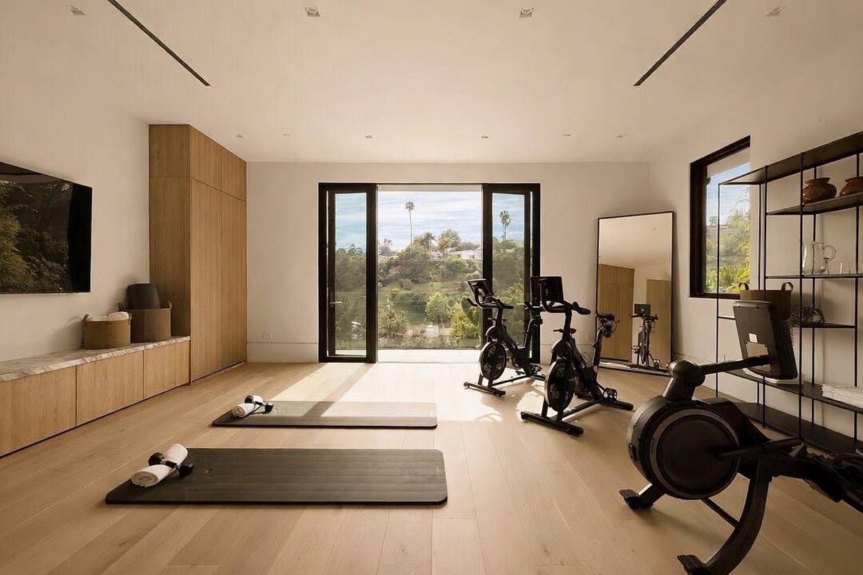 This bright and airy home gym features light wood flooring and minimalist white walls, creating a clean, professional workout environment. The space is equipped with two stationary bikes, a rowing machine, and yoga mats, while a large mirror and floor-to-ceiling glass doors provide an expansive feel and a scenic view of the surrounding hills. A long, low-profile storage unit with light wood cabinetry runs along the left wall, offering functional organization for fitness equipment.