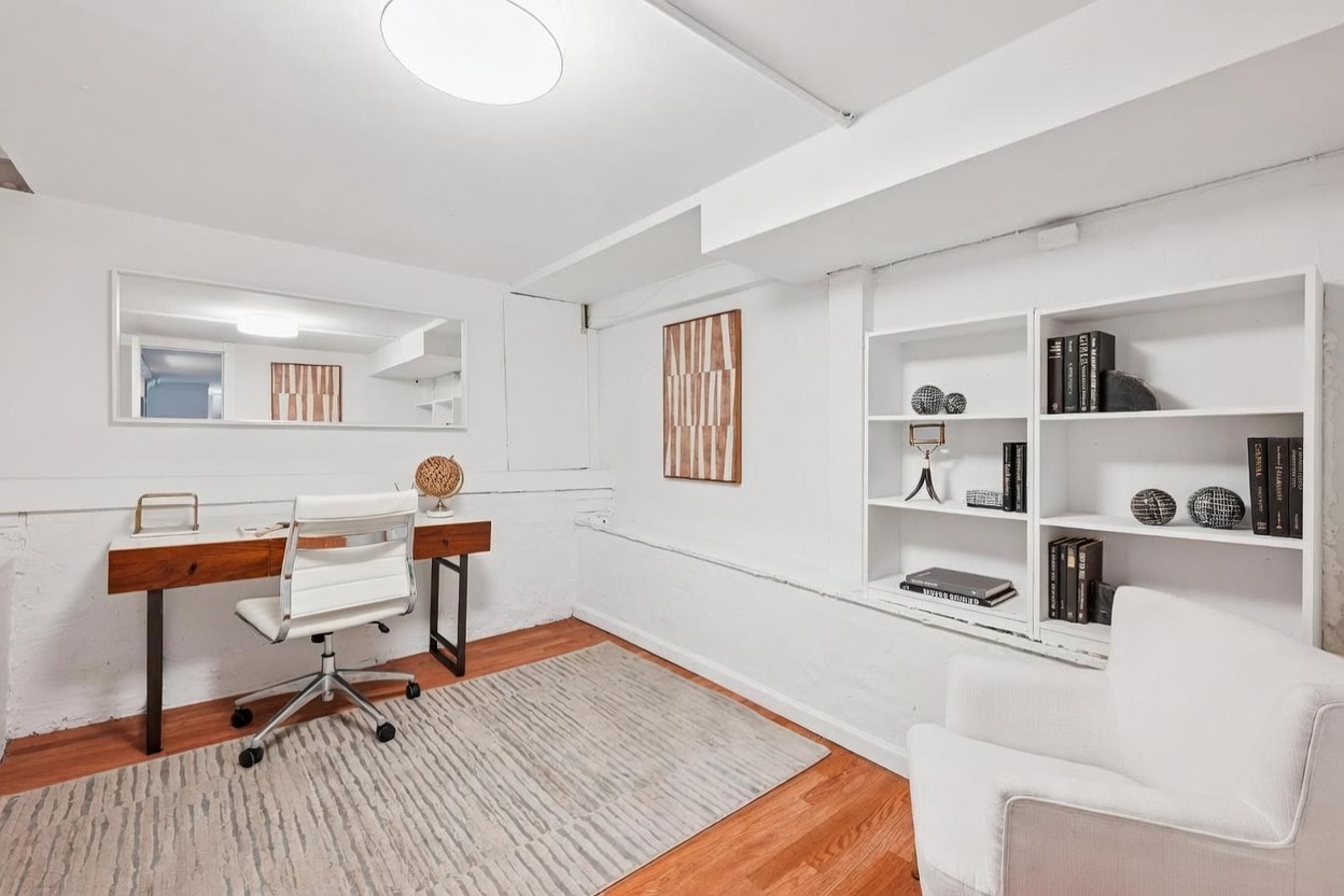 This bright and minimalist home office features white-painted brick walls, warm wood flooring, and a neutral-toned area rug. The space is furnished with a sleek, light-wood desk paired with a white ergonomic chair, complemented by built-in white shelving units and a comfortable armchair in the foreground. A large horizontal mirror above the desk enhances the sense of space, creating a clean and productive atmosphere.