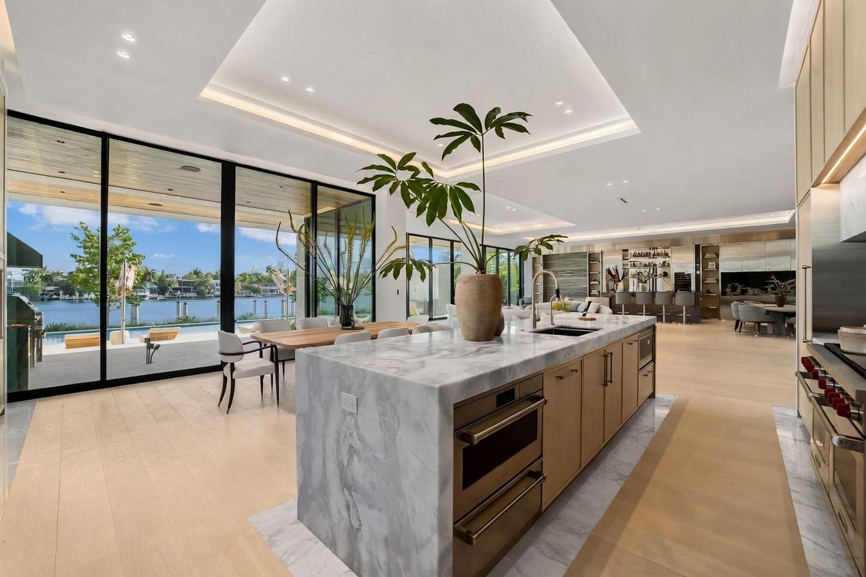 This expansive, open-concept kitchen features a large stone-topped island with integrated appliances and light wood cabinetry. The space flows seamlessly into a dining area and living room, all framed by floor-to-ceiling glass walls that provide a panoramic view of the waterfront and pool area. The design emphasizes a bright, airy atmosphere with recessed lighting and a neutral color palette, creating a sophisticated and luxurious living environment.