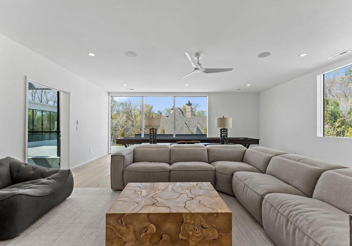 This spacious, modern living room features a large, neutral-toned sectional sofa arranged around a unique, organic-shaped wooden coffee table. The room is bathed in natural light from expansive windows that offer a view of the outdoors, while a pool table sits in the background near the glass. The minimalist aesthetic is complemented by light wood flooring and a clean, white color palette, creating an airy and inviting atmosphere.