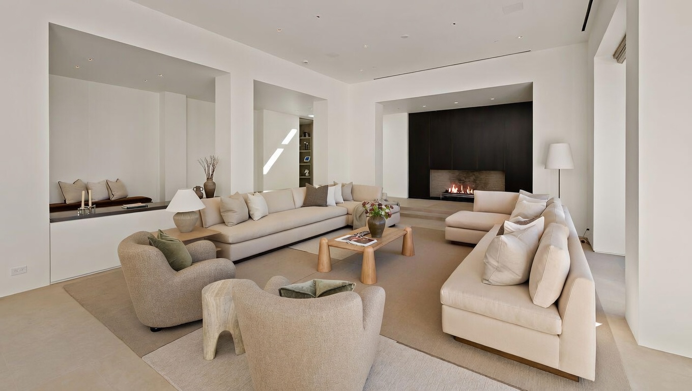 This spacious and minimalist living room features a neutral color palette with plush, cream-colored sectional sofas and rounded armchairs arranged around a light wood coffee table. A striking dark accent wall with a built-in linear fireplace serves as the focal point, while recessed lighting and clean lines enhance the modern, airy atmosphere. The perspective is wide-angle, capturing the expansive layout and the seamless integration of the seating area with the surrounding architectural features.
