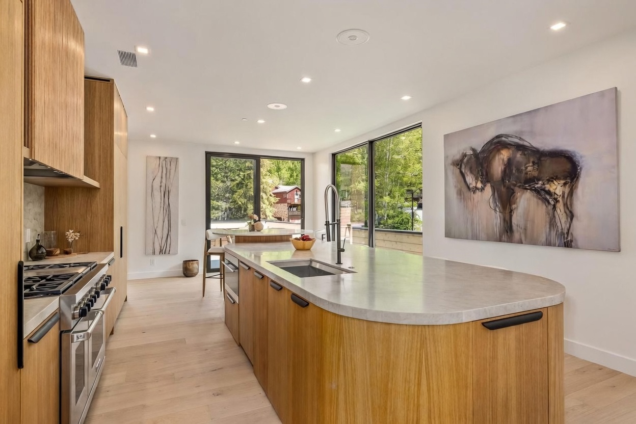 This modern kitchen features a large, curved island with a stone countertop and light wood cabinetry, providing a sleek and contemporary aesthetic. The space is illuminated by recessed lighting and large windows that offer a view of the surrounding greenery. A prominent piece of abstract art hangs on the wall, adding a sophisticated touch to the bright, open-concept design.