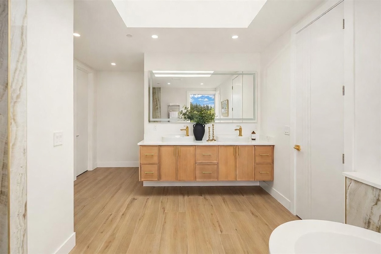 This bright and airy primary bathroom features a double vanity with light wood cabinetry and gold-toned hardware, topped with a clean white surface. A large, rectangular mirror spans the wall above the sinks, reflecting a window that brings in natural light, while a skylight overhead enhances the open feel. The space is finished with light-toned wood-look flooring and crisp white walls, creating a modern and minimalist aesthetic.