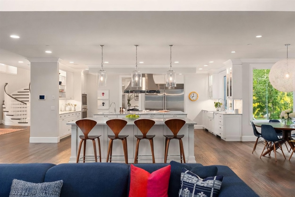 This bright, open-concept kitchen features a large central island with four wooden bar stools, white cabinetry, and stainless steel appliances. The space is illuminated by three glass pendant lights and recessed lighting, with a view of a curved staircase to the left and a dining area to the right. The warm wood flooring and clean, neutral color palette create an inviting and sophisticated atmosphere.