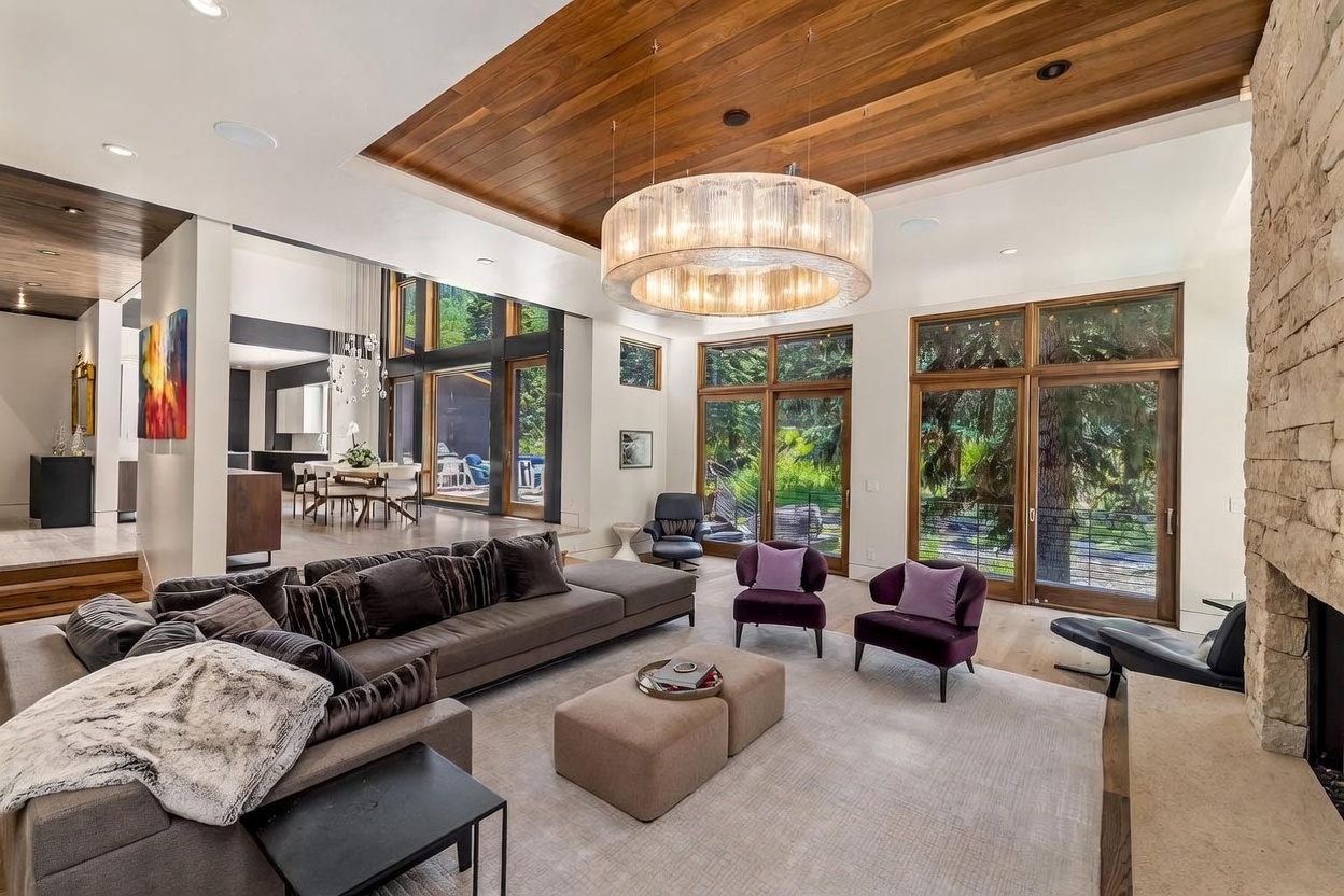 This expansive living room features a high wood-paneled ceiling with a large, circular crystal chandelier as a central focal point. The space is anchored by a deep, neutral-toned sectional sofa and two vibrant purple accent chairs, all arranged around a pair of rectangular ottomans on a light-colored area rug. A prominent stone-clad chimney breast rises on the right, while large windows provide an abundance of natural light and views of the surrounding trees.