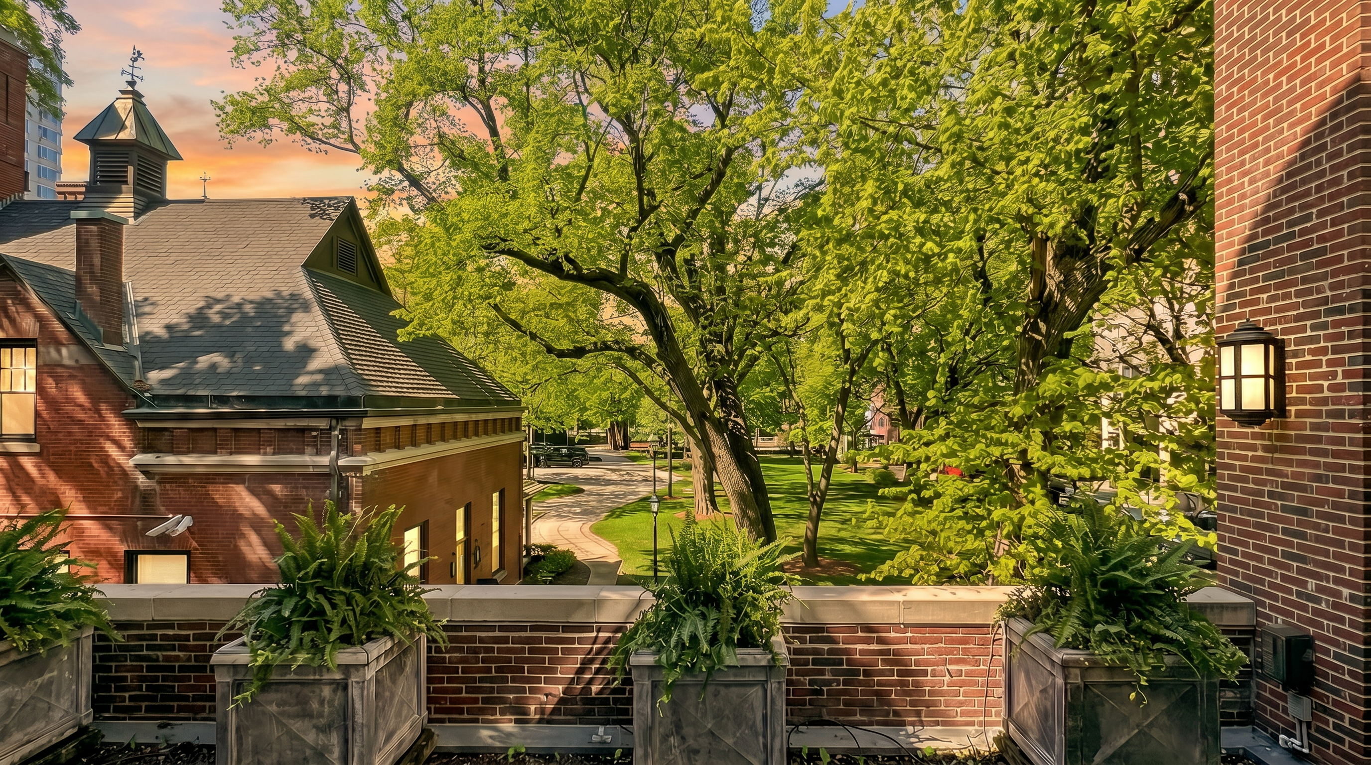 This elevated view captures a private terrace featuring stone flooring and three large, weathered planters filled with lush greenery. The vantage point overlooks a serene, tree-lined park area with a brick building to the left and a modern brick-clad structure to the right. The scene offers a peaceful, urban-oasis atmosphere with clear blue skies above.