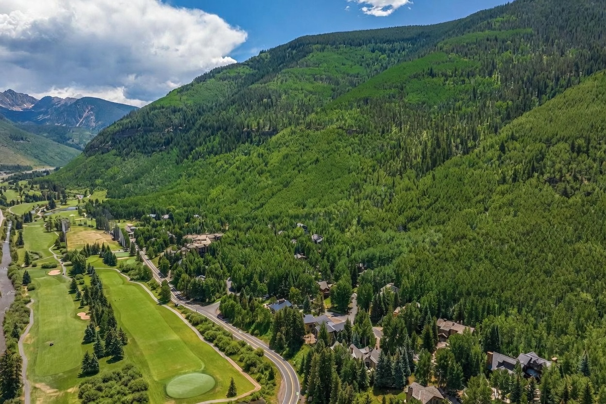 This high-angle aerial view captures a lush, mountainous landscape featuring a winding road that borders a manicured golf course. Nestled among the dense forest are several luxury homes, showcasing a serene and secluded residential setting. The expansive backdrop of towering, tree-covered mountains creates a dramatic and picturesque environment.