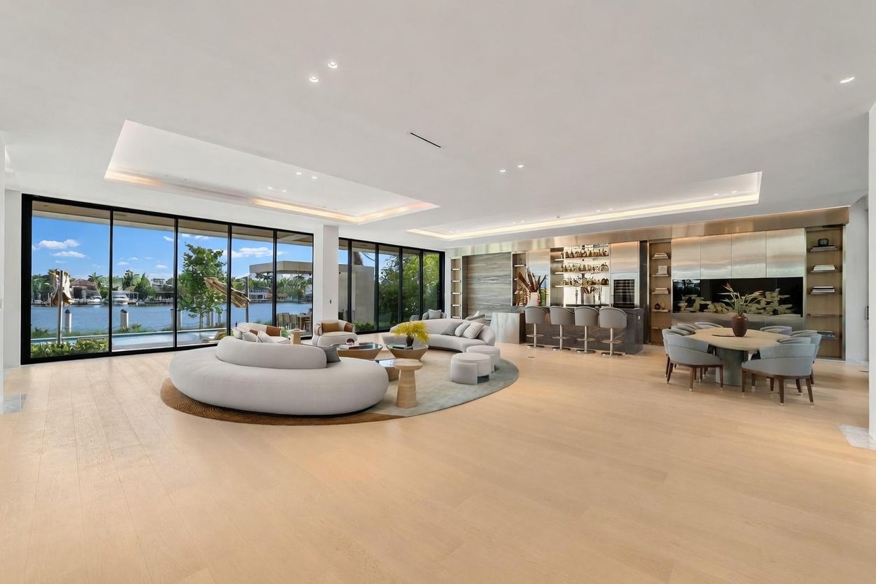 This expansive, open-concept living area features light wood flooring and a neutral color palette, creating a bright and airy atmosphere. The space is anchored by a large, curved sofa set on a circular rug, with a dining area and a sleek bar located in the background. Massive floor-to-ceiling windows provide panoramic views of the waterfront, seamlessly blending the indoor living space with the outdoor environment.