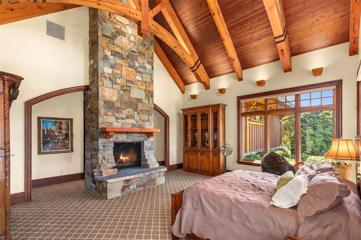 This spacious primary bedroom features a vaulted ceiling with exposed timber beams and a warm wood-paneled finish. A prominent stone-clad chimney breast serves as a central focal point, complemented by a large window that invites natural light into the room. The space is furnished with a comfortable bed and a classic wooden cabinet, creating a cozy and rustic atmosphere.