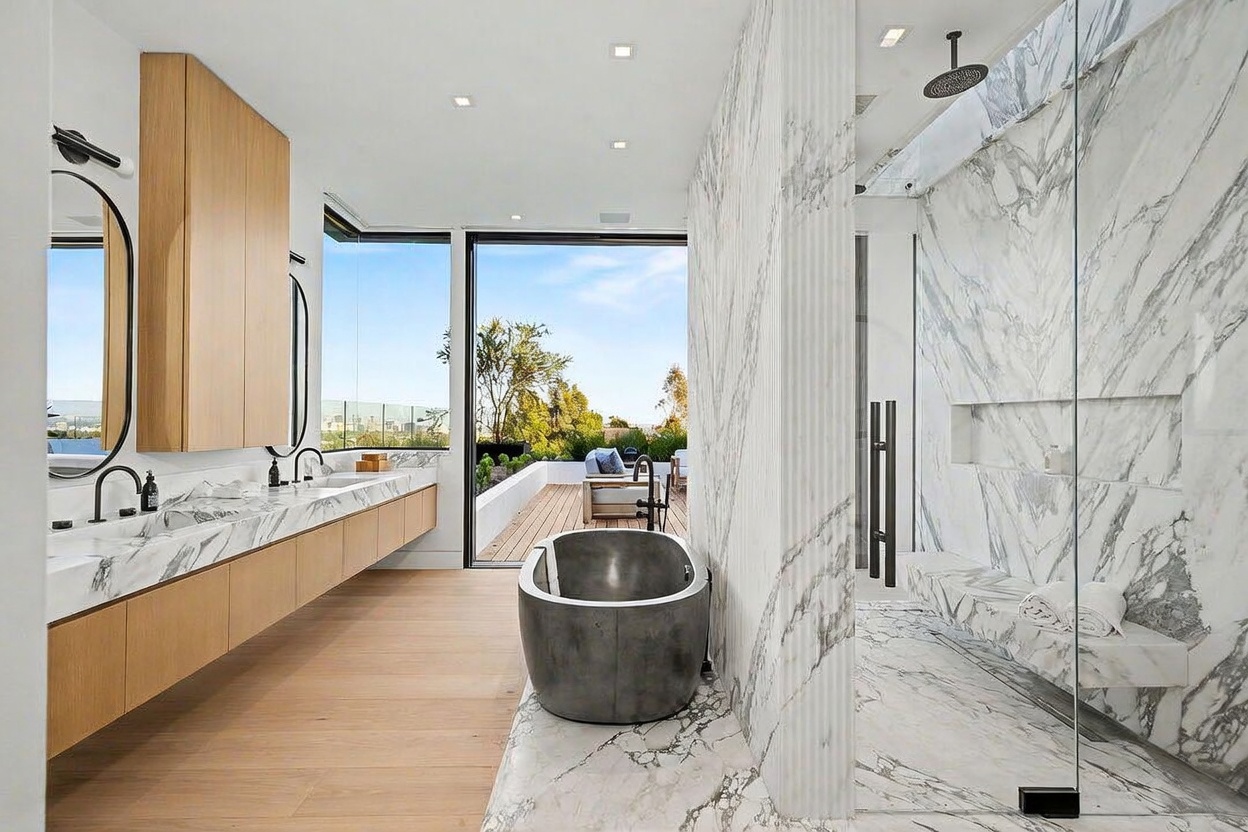This luxurious primary bathroom features a sleek, modern design with a floating vanity topped with light-veined stone and a deep, sculptural soaking tub. A glass-enclosed shower area with a built-in stone bench sits adjacent to a large sliding glass door that opens onto an outdoor deck. The space is bathed in natural light, creating a seamless transition between the indoor sanctuary and the exterior landscape.