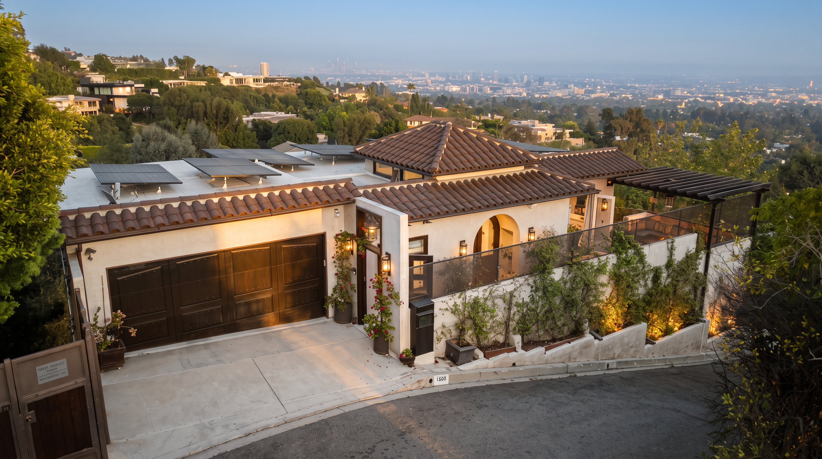 This elevated perspective captures a Mediterranean-style home nestled on a hillside, showcasing a multi-level roofline with dark clay tiles and integrated solar panels. The property features a prominent garage, a gated entryway, and a terraced patio area adorned with climbing greenery and warm exterior lighting. The background offers a sweeping, panoramic view of a sprawling city skyline under a clear twilight sky.