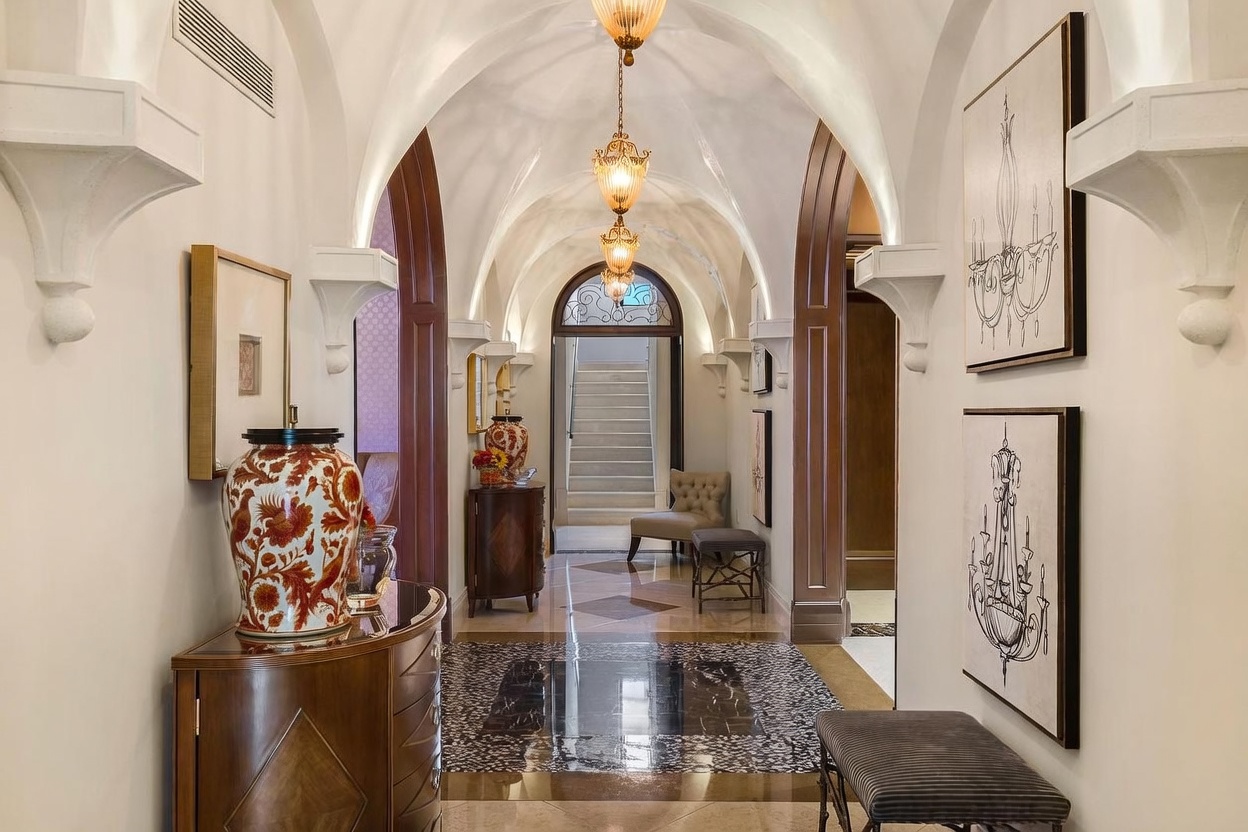 This elegant, arched hallway features a sophisticated Mediterranean-inspired design with high, vaulted ceilings and ornate hanging pendant lights. The space is anchored by a polished marble floor with a decorative inlay, leading the eye toward a grand staircase visible through a glass-paned arched doorway. Framed artwork, a decorative vase on a console table, and a small seating area create a refined and welcoming atmosphere.