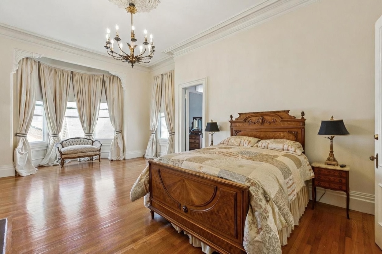 This spacious primary bedroom features polished hardwood floors and a classic, elegant aesthetic. A large, ornate wooden bed frame serves as the centerpiece, complemented by matching bedside tables with lamps and a decorative settee positioned near the tall, draped windows. The room is illuminated by a traditional chandelier, creating a sophisticated and airy atmosphere.