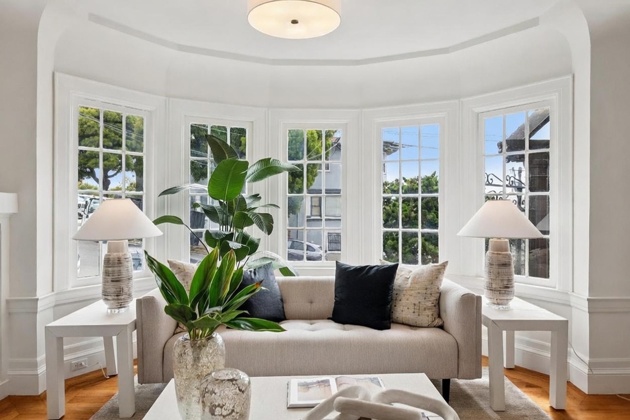 This bright and inviting living room features a curved bay window arrangement that floods the space with natural light. A neutral-toned sofa is centered in front of the windows, flanked by two matching side tables with textured lamps and a large potted plant. The room is finished with warm wood flooring and a clean, minimalist aesthetic that emphasizes the architectural character of the window nook.