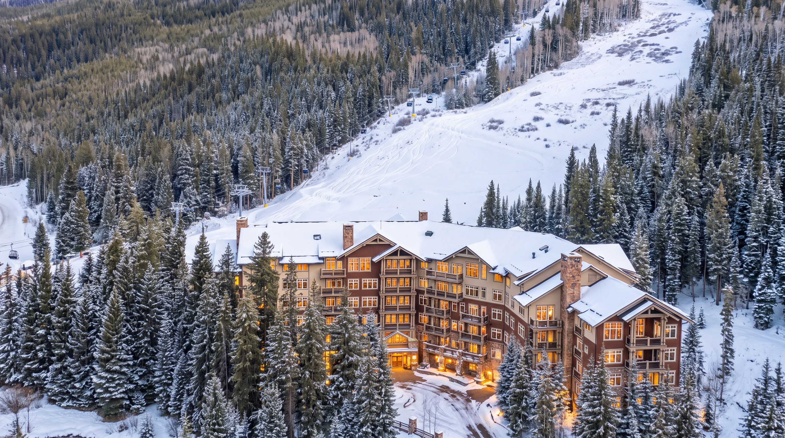 This high-angle aerial shot captures a grand, multi-story mountain lodge nestled within a dense evergreen forest at the base of a ski slope. The architecture features a rustic, lodge-style aesthetic with dark shingled roofs, stone accents, and numerous balconies overlooking the surrounding wilderness. The perspective emphasizes the property's prime location, showcasing the ski lift infrastructure ascending the mountain directly behind the building.