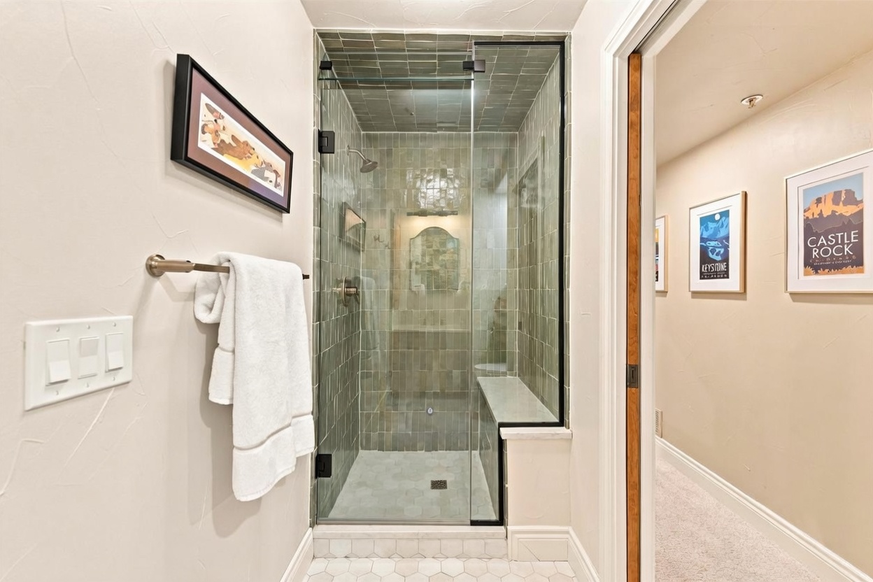 This bathroom features a walk-in shower with a glass enclosure, framed in black, and lined with sage-green rectangular tiles. The shower includes a built-in stone bench and a hexagonal tile floor that extends into the adjacent hallway. To the right, a doorway leads into a carpeted hallway decorated with framed travel posters, creating a cohesive and inviting transition between spaces.