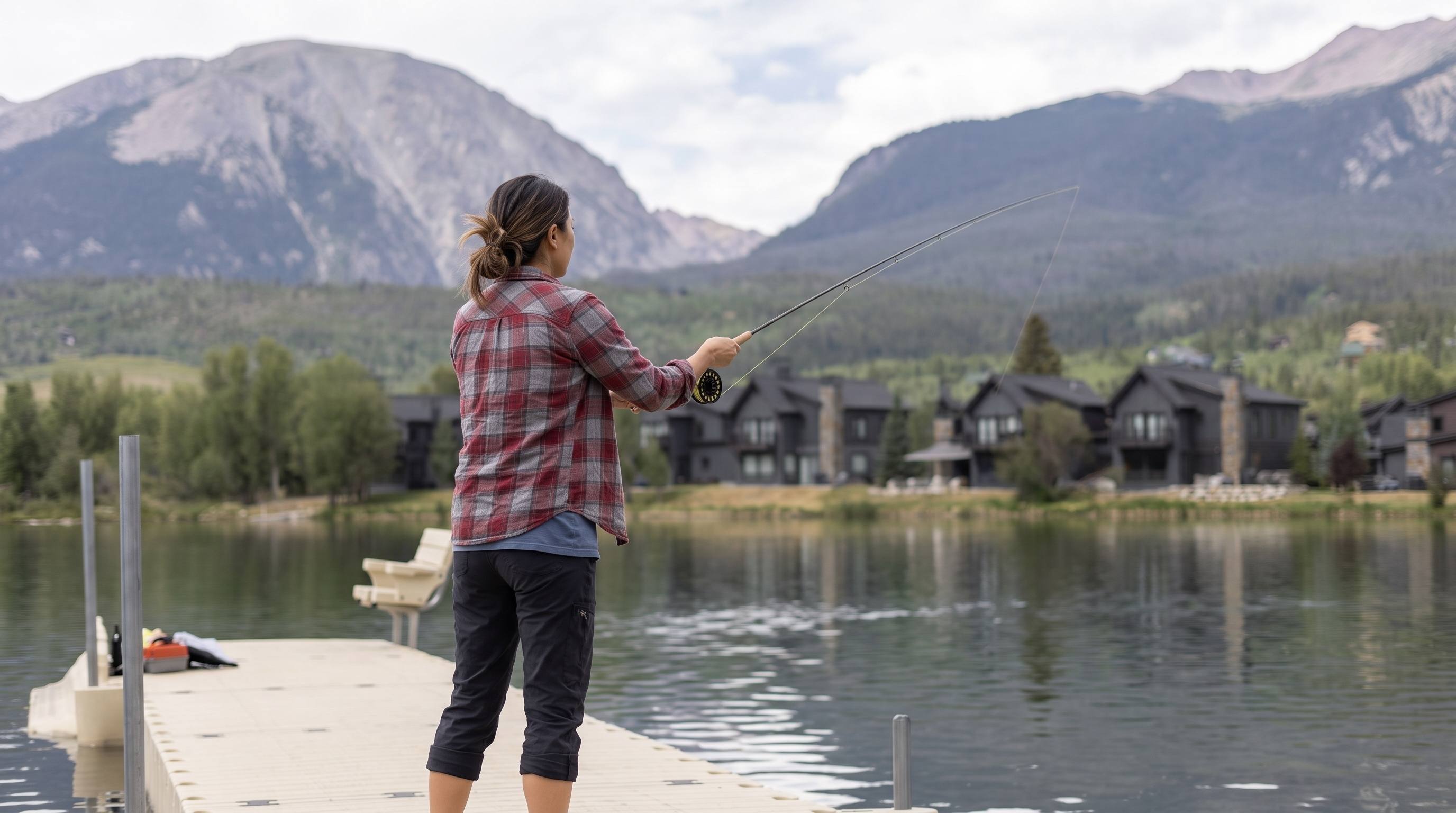 This scenic lakefront scene features a floating dock utilized for recreational fishing, set against a backdrop of luxury mountain residences. The composition highlights a peaceful community amenity with panoramic views of rugged peaks and lush greenery. It conveys an active, upscale lifestyle opportunity in a highly desirable natural setting.