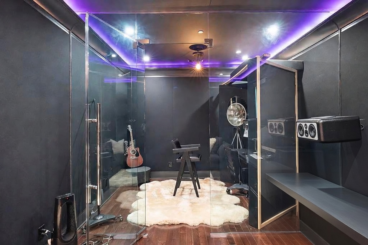 This is an interior shot of a modern recording studio. The room features soundproofing panels, glass partitions, and professional recording equipment. The space is illuminated with purple accent lighting, creating a contemporary and creative atmosphere.
