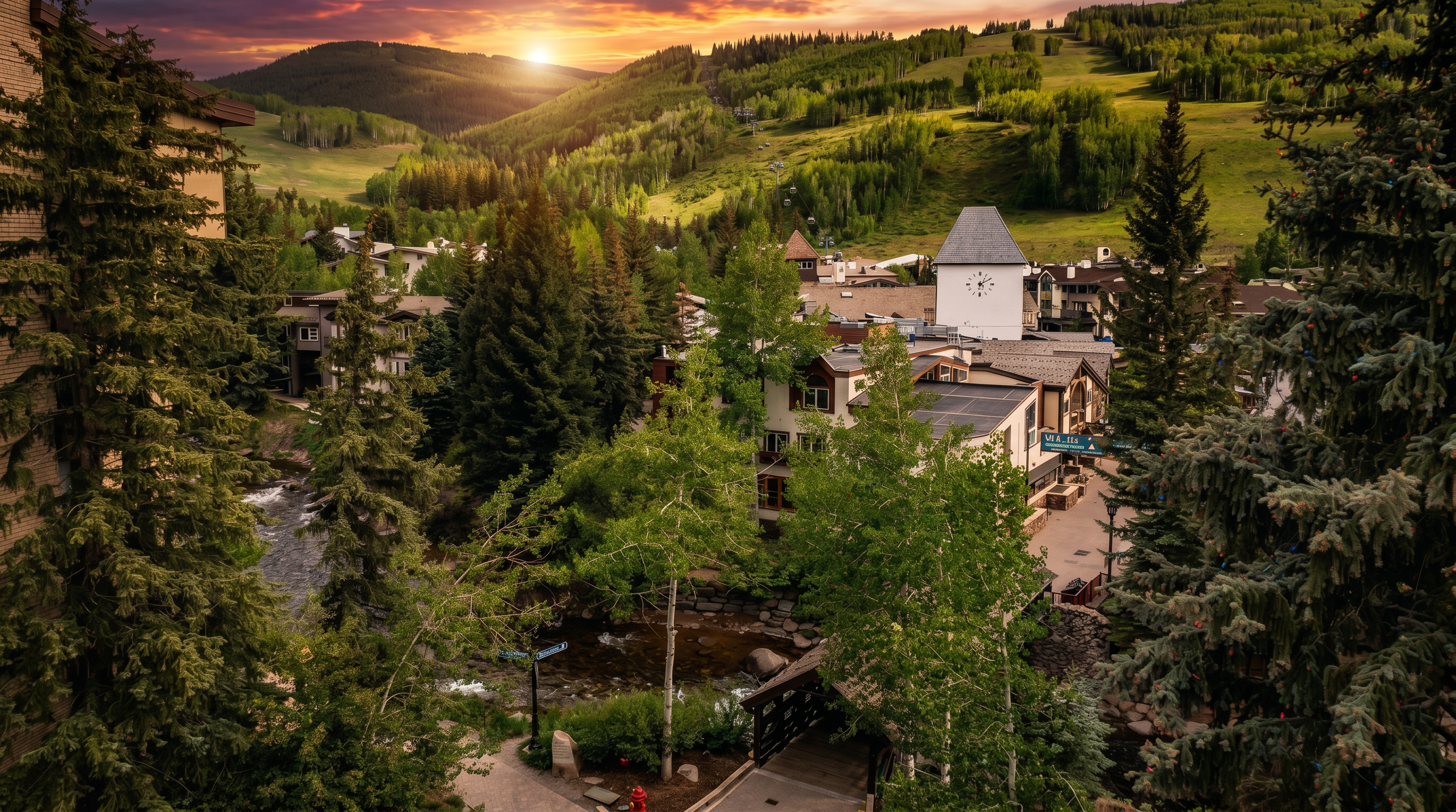 This elevated perspective captures a scenic mountain village nestled at the base of a lush, green ski slope. The scene features a winding creek flowing past a wooden footbridge, surrounded by dense evergreen and aspen trees, with a prominent clock tower serving as a central landmark. The architecture reflects a classic alpine style, blending harmoniously with the natural landscape under a bright, partly cloudy sky.