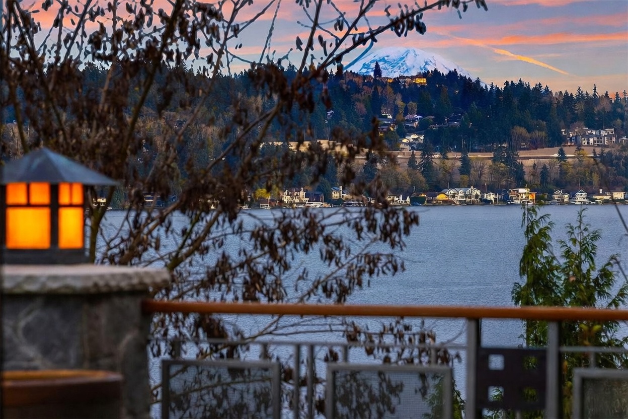 This image captures a serene, elevated view from a balcony overlooking a tranquil lake with a majestic, snow-capped mountain in the distance. In the foreground, a warm, glowing outdoor lamp sits on a stone pillar, framing the scene alongside the silhouettes of tree branches. The composition emphasizes a peaceful, high-end lifestyle, highlighting the scenic waterfront location and the beautiful sunset sky.