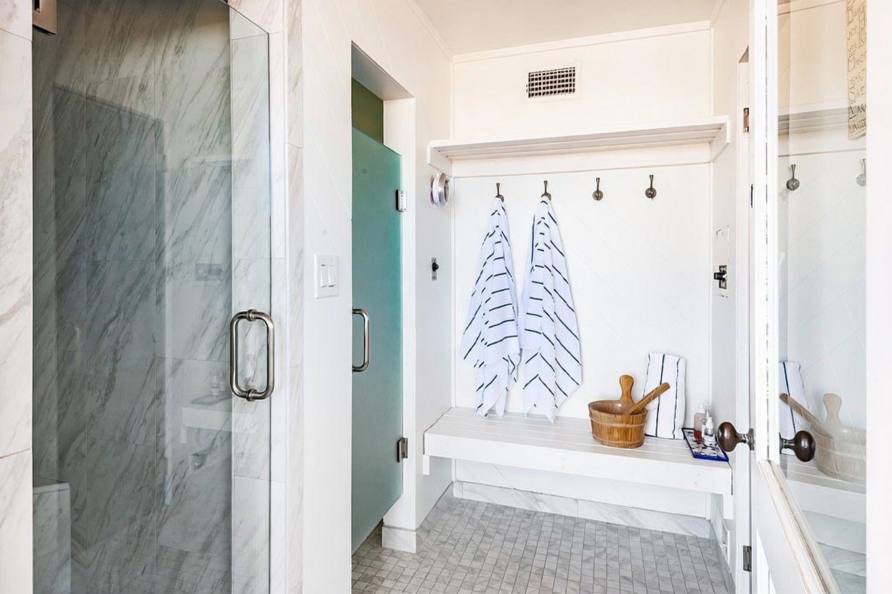 This bright, transitional-style bathroom features a walk-in shower with light-toned stone tile walls and a glass door. Adjacent to the shower is a built-in bench area with hooks for towels, providing a functional mudroom-style transition space. The floor is covered in small, neutral-toned mosaic tiles, and the overall aesthetic is clean, airy, and well-organized.