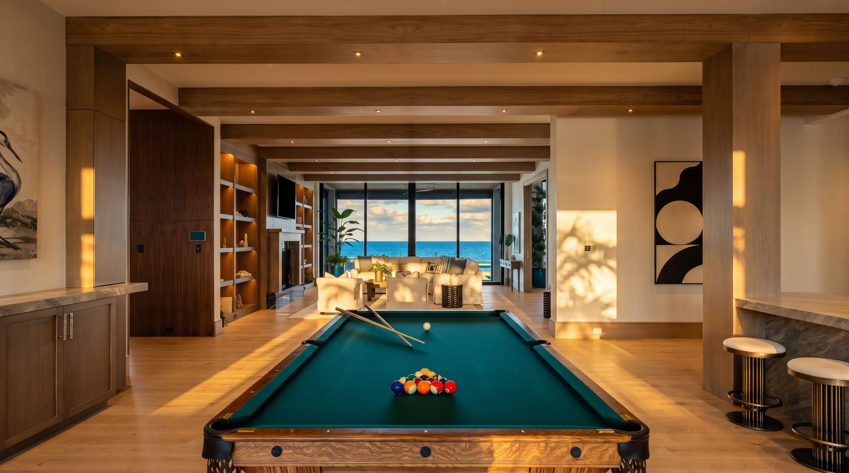 This expansive open-concept living space features a prominent pool table in the foreground, leading the eye toward a bright seating area with a direct view of the ocean. The room is defined by warm wood-toned ceiling beams and light wood flooring, creating a cohesive and airy atmosphere. Large glass panels at the rear provide an unobstructed connection to the coastal scenery, while built-in shelving and a fireplace add architectural interest to the living area.