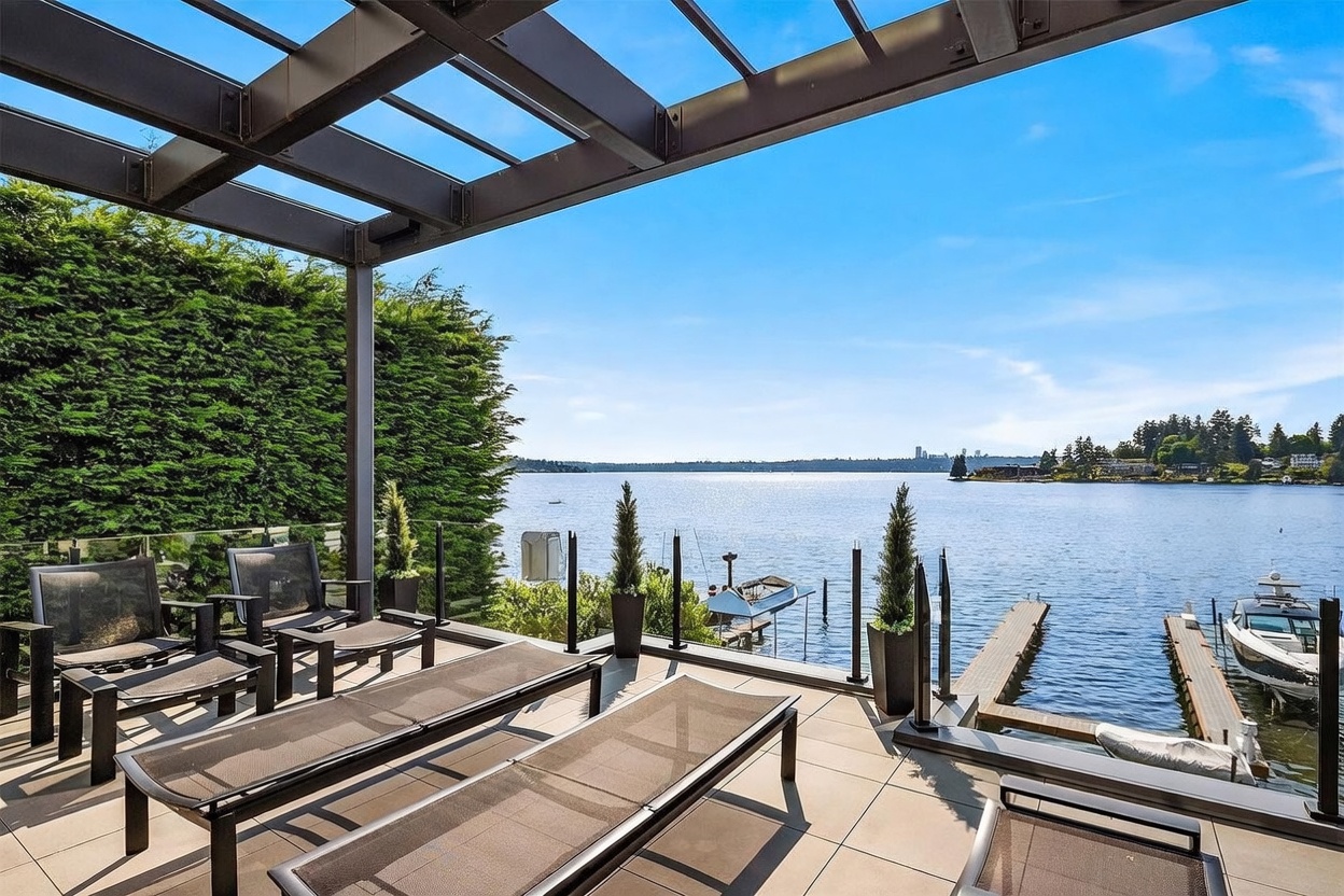 This elevated deck offers a stunning, unobstructed view of a calm lake with a distant city skyline visible on the horizon. The space is furnished with modern lounge chairs and features a sleek metal pergola structure, glass railings, and potted greenery that frame the waterfront scenery. A private dock with a boat is visible below, emphasizing the luxurious, resort-like lifestyle of this waterfront property.