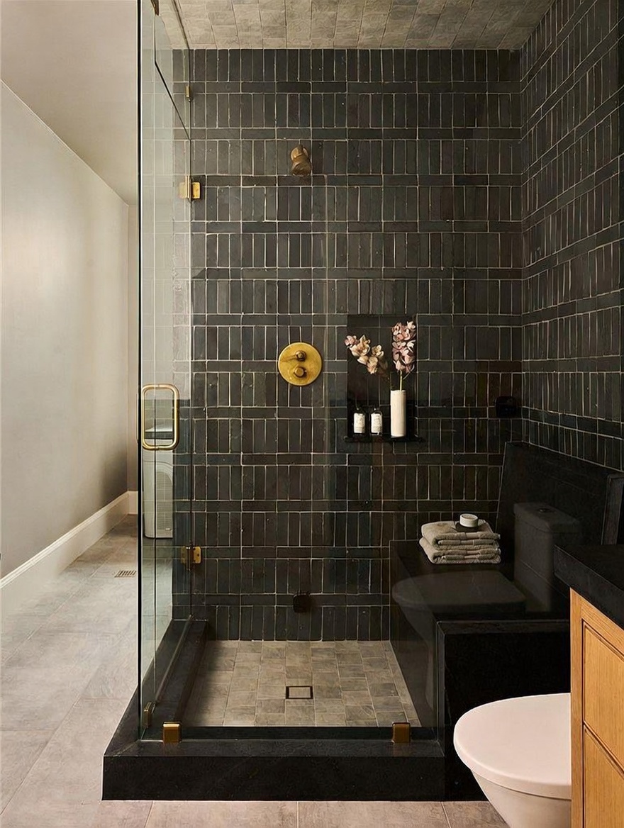 This modern bathroom features a walk-in shower enclosed by glass panels, showcasing dark, rectangular wall tiles and a contrasting stone-tiled floor. A built-in bench inside the shower provides convenience, while gold-toned fixtures add a touch of luxury against the moody color palette. The space is accented by a small niche containing decorative flowers and toiletries, creating a sophisticated and spa-like atmosphere.