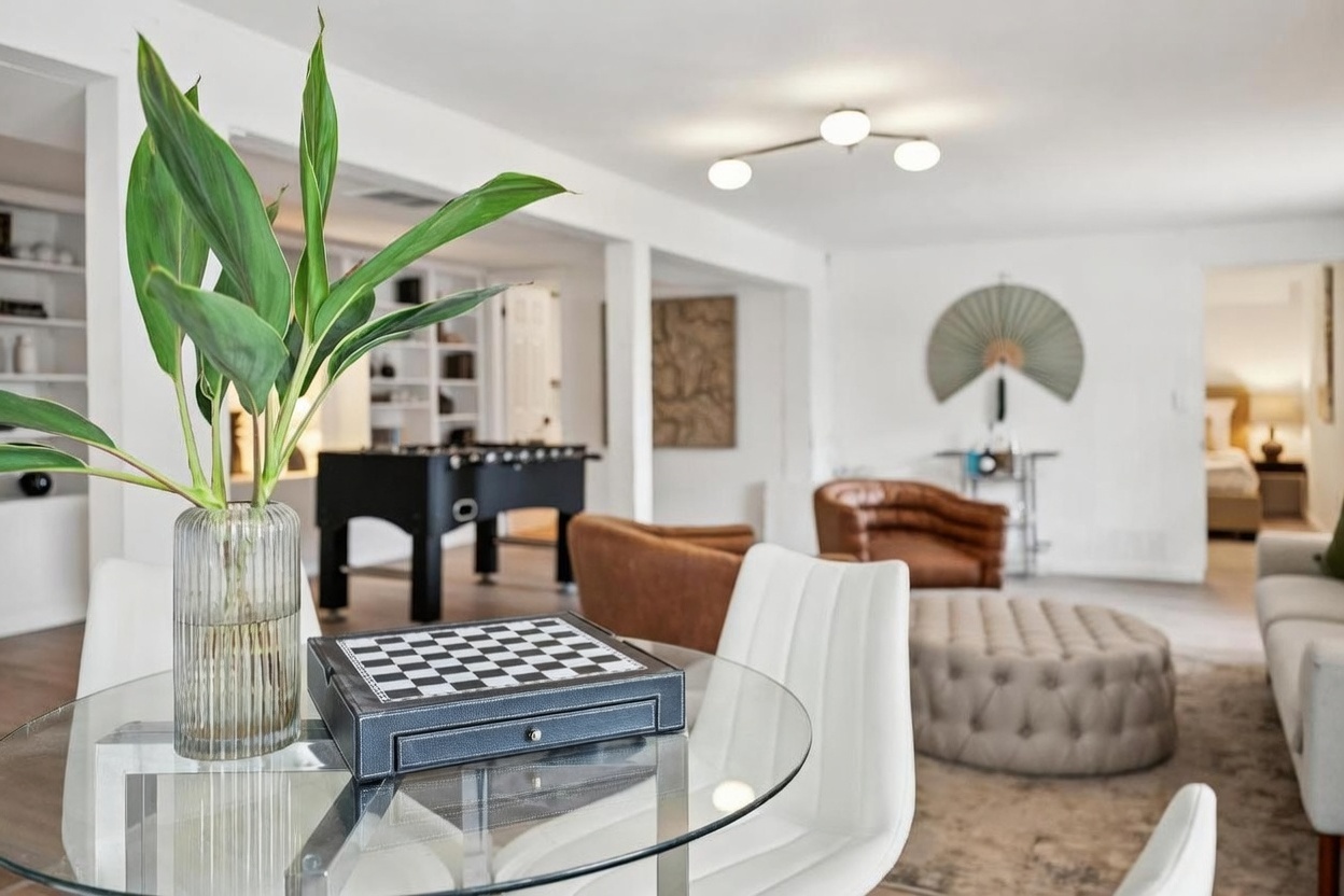 This bright, open-concept living space features a foreground dining area with a glass-topped table, a decorative chessboard, and a vibrant green plant in a ribbed glass vase. The background reveals a spacious living area furnished with brown leather armchairs, a tufted ottoman, and a foosball table, creating a versatile and inviting atmosphere. The room is illuminated by a modern, multi-arm light fixture and maintains a clean, contemporary aesthetic with white walls and neutral decor.