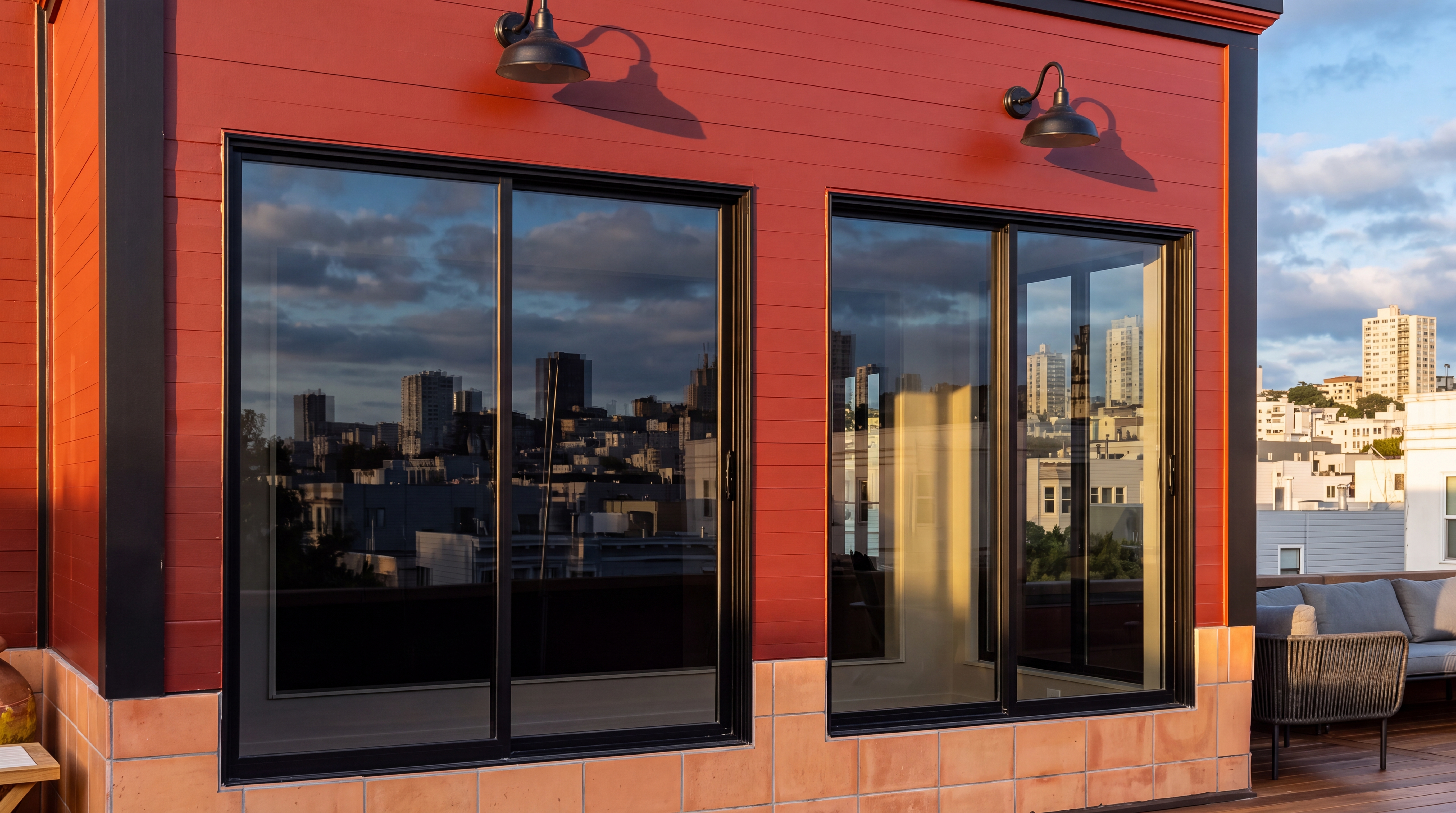 This outdoor terrace features a striking red horizontal paneled siding contrasted by modern black-framed sliding glass doors. The area includes comfortable outdoor sectional seating on a wooden deck, set against a stunning urban skyline backdrop. The space is accessorized with wall-mounted, vintage-style industrial lamps, creating a sophisticated and inviting urban retreat.