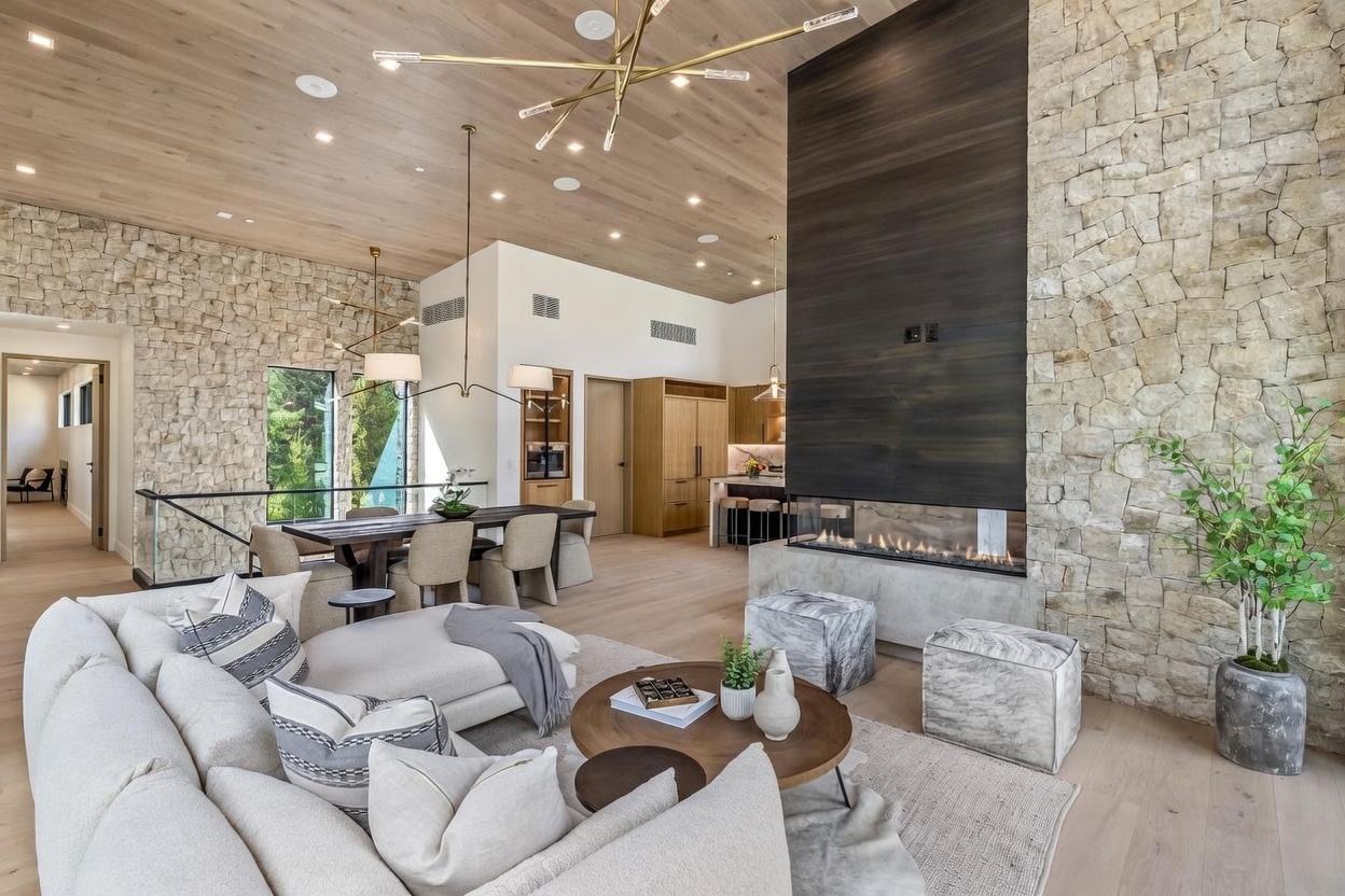 This expansive open-concept living area features a light wood vaulted ceiling and a prominent stone-clad chimney breast that anchors the space. A large, plush sectional sofa faces a modern linear fireplace, while a dining area with a dark wood table sits adjacent to a bright kitchen in the background. The room is bathed in natural light from large windows, creating a warm, sophisticated atmosphere with a blend of natural textures and contemporary design.