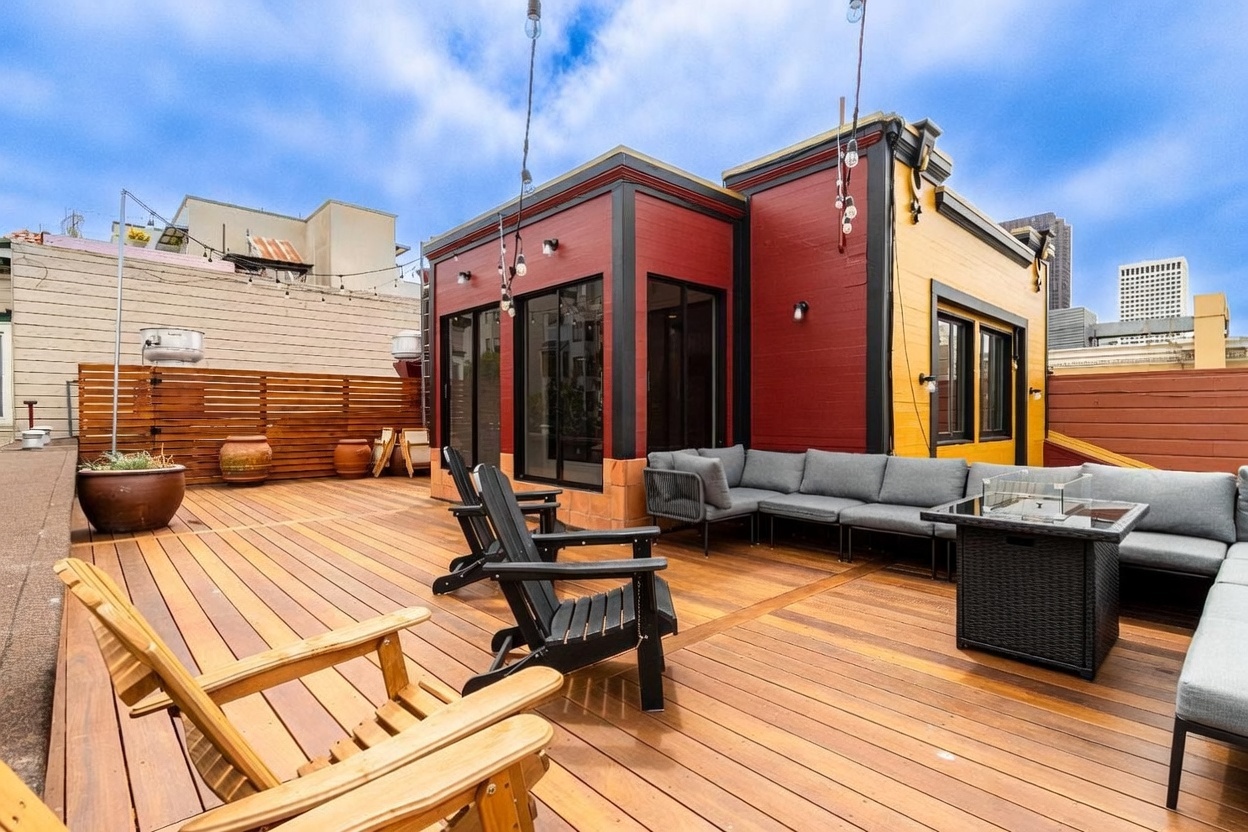 This spacious rooftop deck features warm-toned wood plank flooring and a contemporary seating area arranged around a central fire pit table. The structure showcases a bold color palette with red and yellow exterior walls, complemented by large glass sliding doors that provide seamless indoor-outdoor flow. String lights hang overhead, creating an inviting atmosphere for entertaining against a backdrop of urban skyline views.