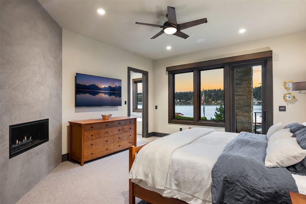 This primary bedroom features a modern aesthetic with a neutral color palette and a prominent fireplace set into a textured, gray-toned wall. A wooden dresser sits beneath a wall-mounted television, while large windows provide a scenic view of the water and surrounding landscape. The room is anchored by a comfortable bed with layered bedding and illuminated by a contemporary ceiling fan.
