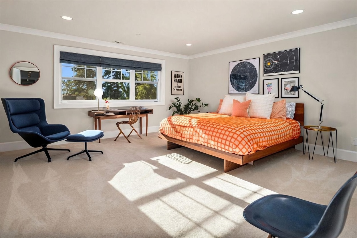 This spacious bedroom features a modern aesthetic with a low-profile wooden bed frame topped with a vibrant orange checkered duvet. A dedicated workspace with a desk and chair is positioned near a large window, while a blue lounge chair and ottoman provide a comfortable seating area. The room is decorated with framed wall art and a potted plant, creating a bright and inviting atmosphere.
