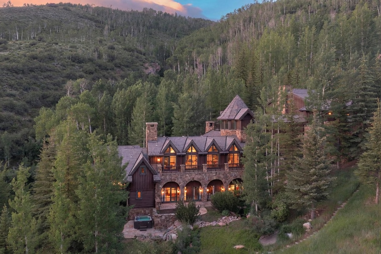 This aerial perspective captures a grand, multi-story mountain estate nestled within a lush forest of aspen and pine trees. The home features a rustic architectural style with stone masonry, dark wood siding, and multiple gabled rooflines, complemented by a private hot tub on the lower stone patio. The warm glow from the interior lighting creates an inviting atmosphere against the backdrop of a twilight sky and dense mountain foliage.