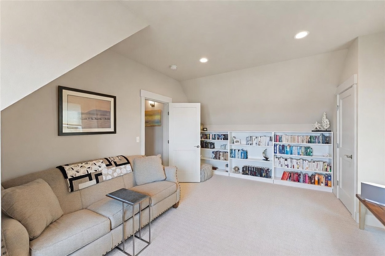 This cozy attic space features a neutral color palette with light-toned carpeting and soft gray walls, creating a serene atmosphere. A comfortable sofa with a patterned throw blanket sits opposite a long, built-in white bookshelf that spans the wall, providing ample storage for books and decor. The room is illuminated by recessed lighting and includes a doorway leading to an adjacent area, making it a versatile living or reading nook.