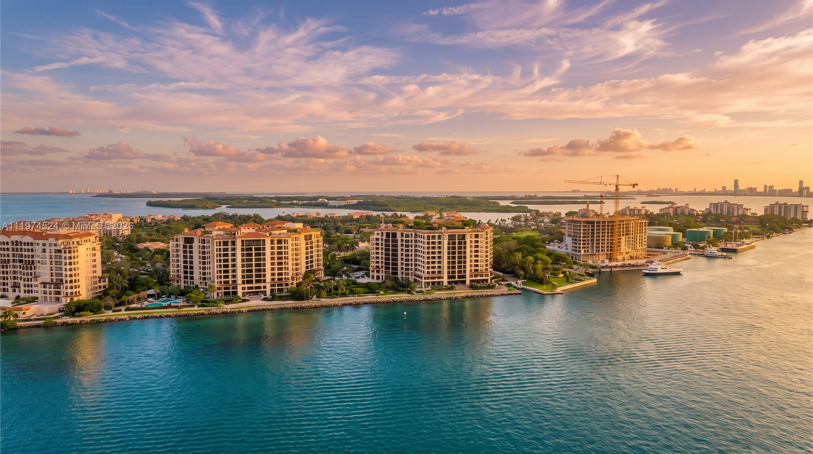 This high-angle aerial shot captures a waterfront residential complex situated on a lush, tropical peninsula surrounded by vibrant turquoise water. The scene features several multi-story apartment buildings with classic architectural details, set amidst manicured green landscaping and palm trees. In the distance, a construction crane is visible near a building under development, while the horizon reveals a clear blue sky and a faint city skyline.