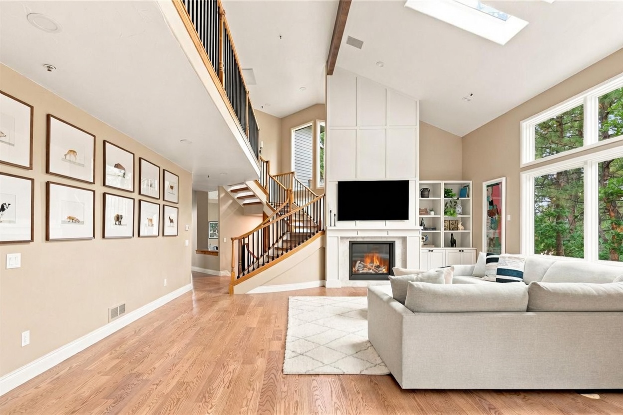 This spacious living room features a light-toned sofa, a patterned area rug, and warm wood flooring. A prominent white fireplace surround with a mounted television serves as the focal point, complemented by built-in shelving and a grand staircase with dark metal railings. Large windows and a skylight fill the room with natural light, creating an airy and inviting atmosphere.
