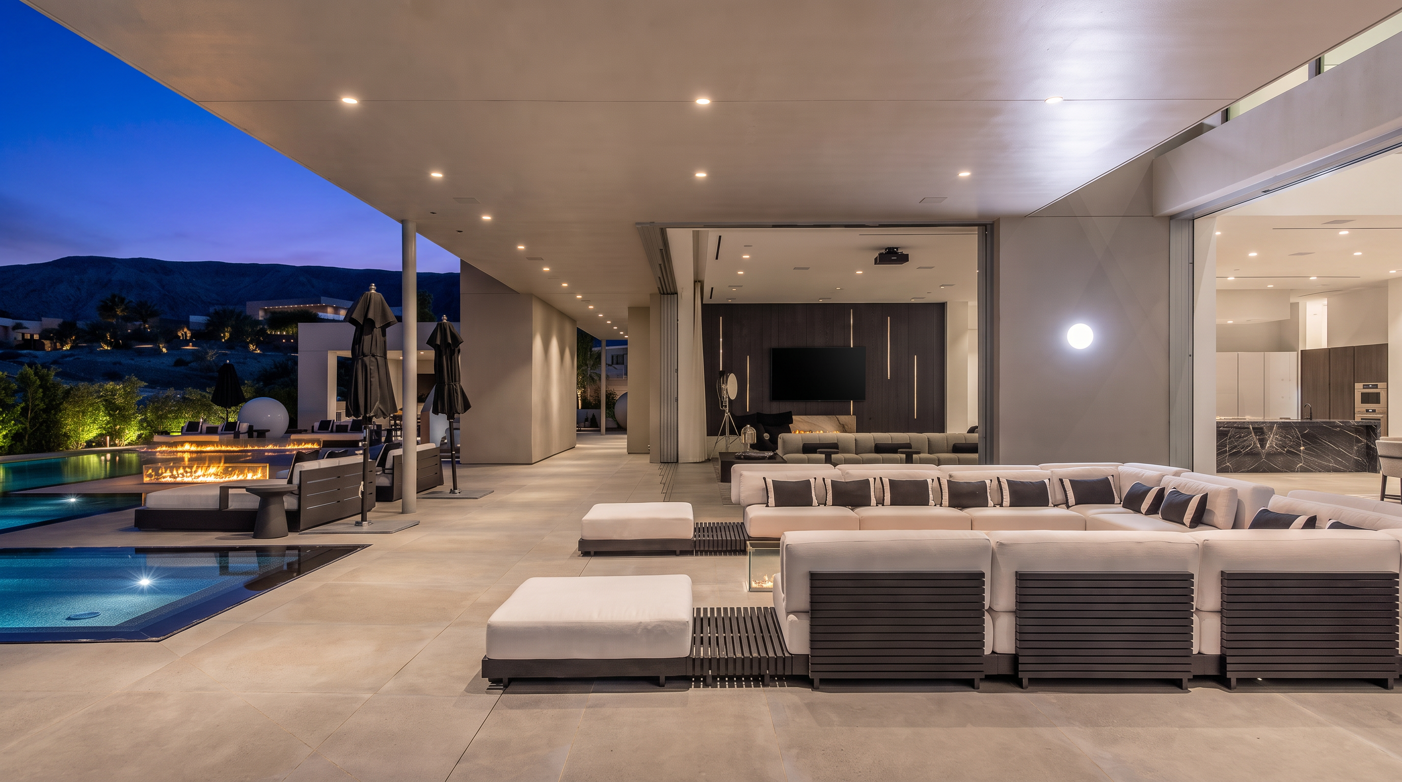 This expansive covered patio seamlessly blends indoor and outdoor living through a wide-open transition to the interior living area. The space features a large, modular white sectional sofa, a sleek fire feature, and a pristine swimming pool that reflects the evening sky. The modern architectural design emphasizes clean lines, recessed lighting, and a sophisticated, resort-like atmosphere perfect for entertaining.